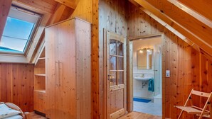 Une pièce en bois avec une lucarne, une armoire, une porte donnant sur une salle de bain et une chaise en bois.