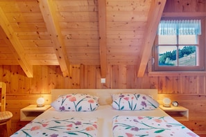 A wooden cabin bedroom with a bed, two bedside tables, and a window with a view of a forest. - Apartment 'Bergblick' with Mountain View, Shared Terrace and Wi-Fi (Mühlenbach)