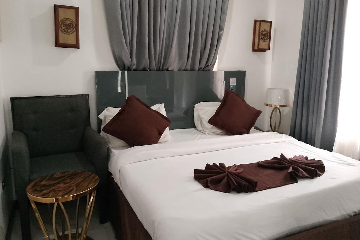 Une chambre d’hôtel avec un lit, deux oreillers, une table de chevet, une chaise, une lampe et des rideaux.