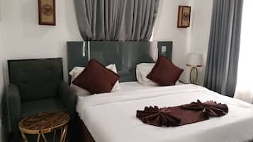 A hotel room with a bed, two pillows, a bedside table, a chair, a lamp, and curtains.