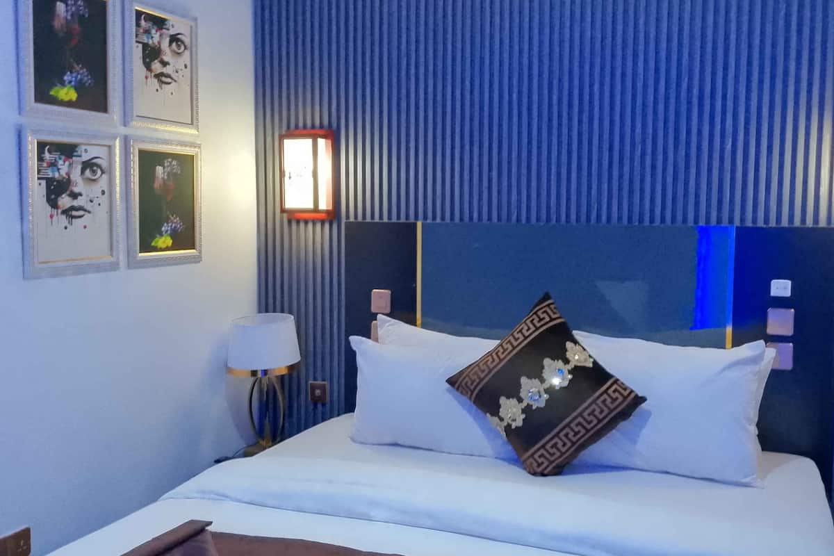 Une chambre d’hôtel avec un grand lit, une table de chevet et une lampe. Des tableaux encadrés ornent le mur.