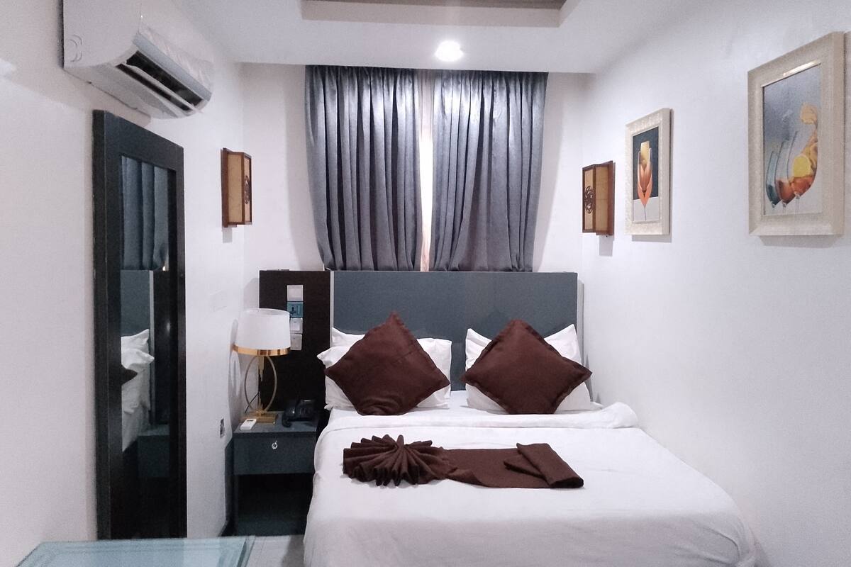 Une chambre d’hôtel avec un lit, une table de chevet, une lampe, un miroir et une fenêtre avec des rideaux.