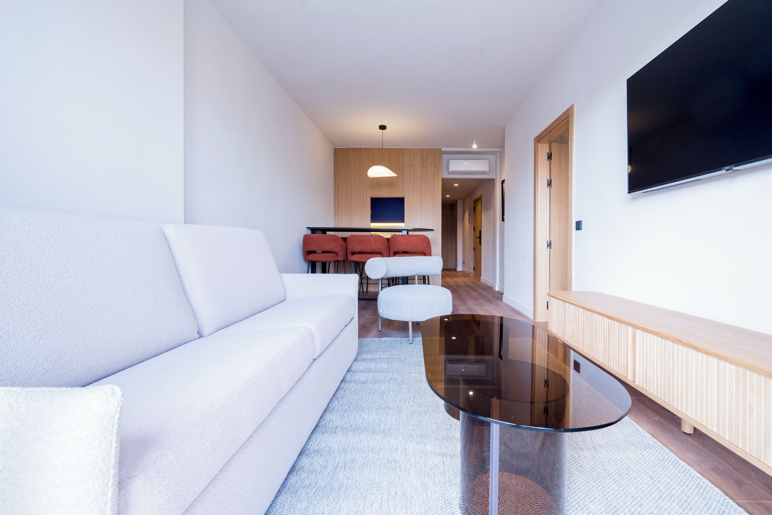 A modern living room with a white sofa, a glass coffee table, and a dining area with red chairs.