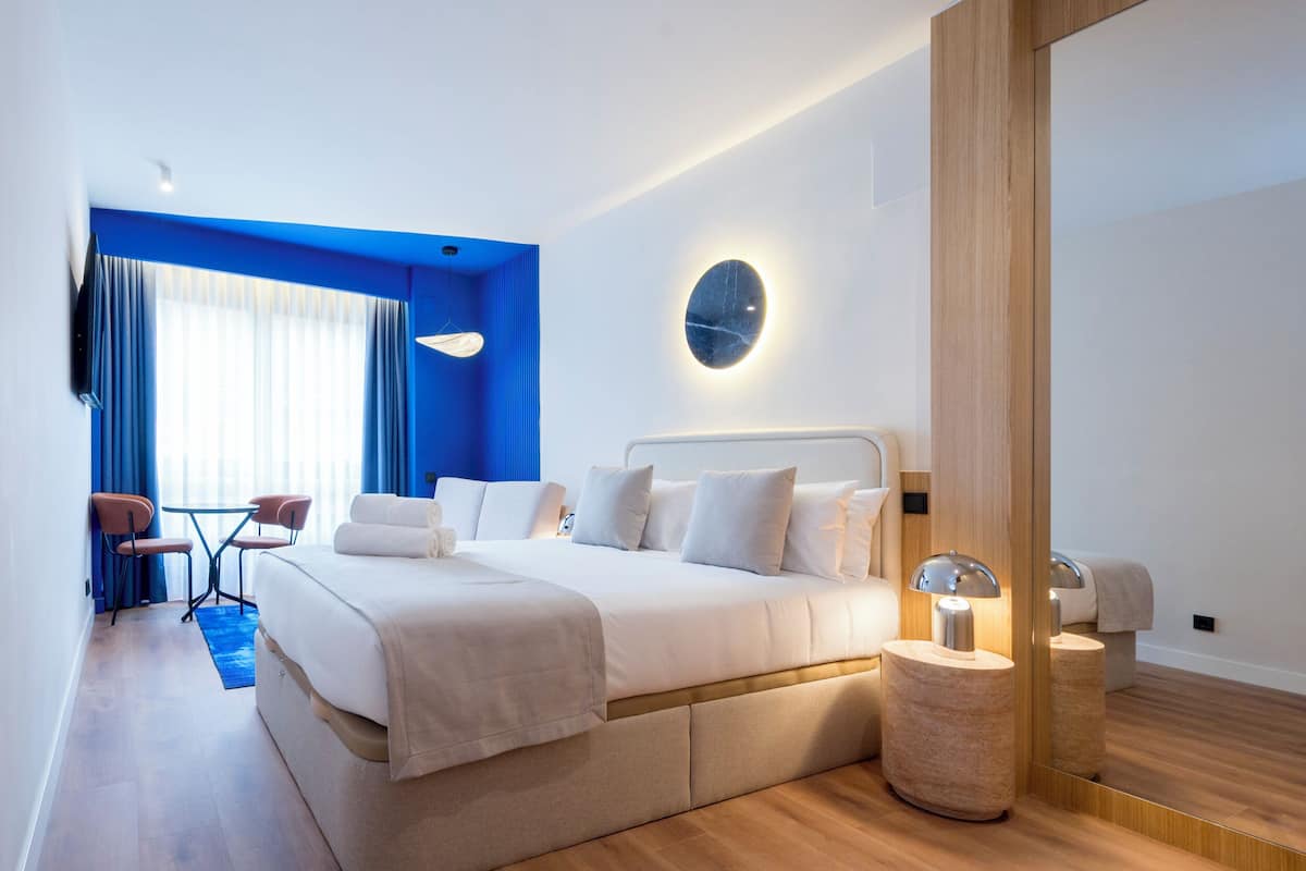 A modern hotel room with a large bed, bedside tables, a mirror, and a seating area with chairs and a table.
