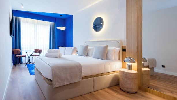 Quarto de hotel moderno com uma cama grande, mesas de cabeceira, um espelho e um espaço de estar com cadeiras e uma mesa.