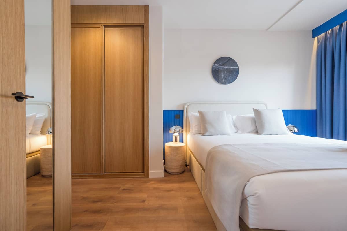 A hotel room with a wooden wardrobe, a round wall clock, a bed with white bedding, and a bedside lamp.