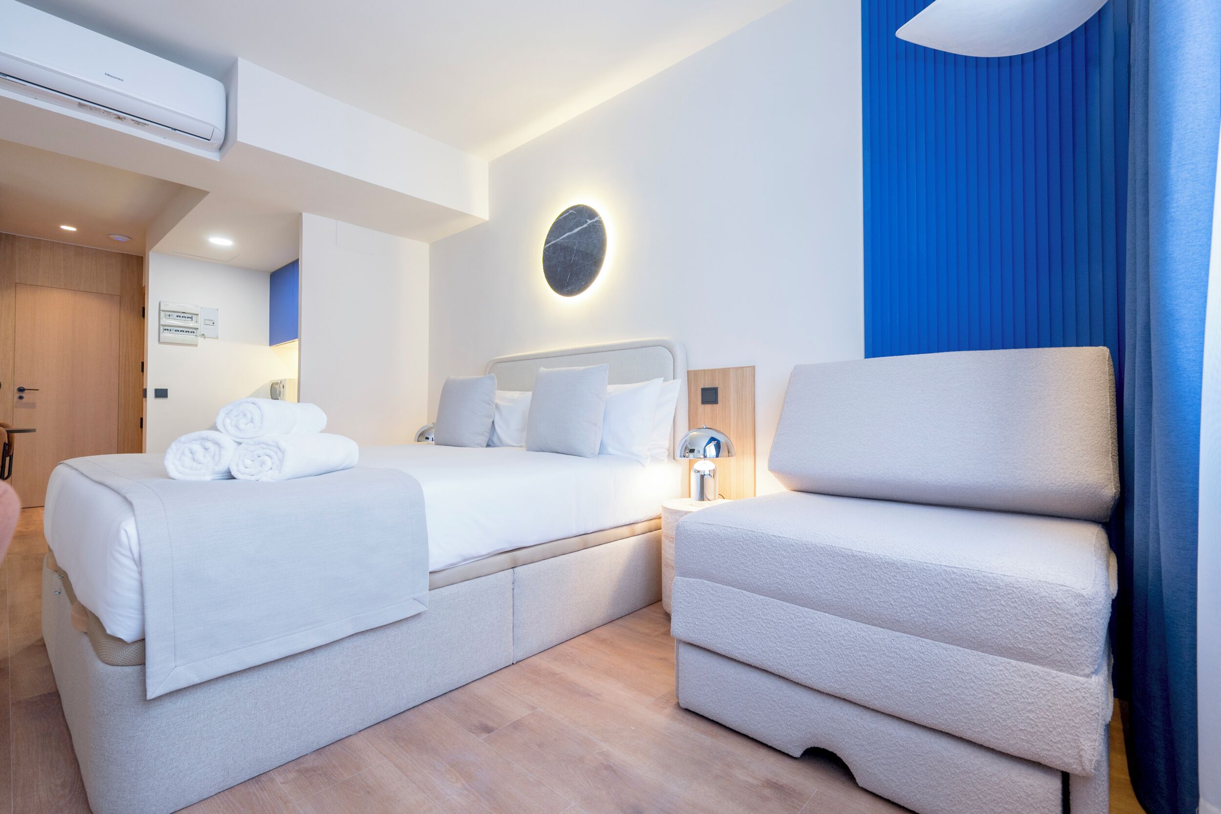 Una habitación de hotel moderna con dos camas, una mesita de noche, una lámpara y un ventanal grande con cortinas azules.
