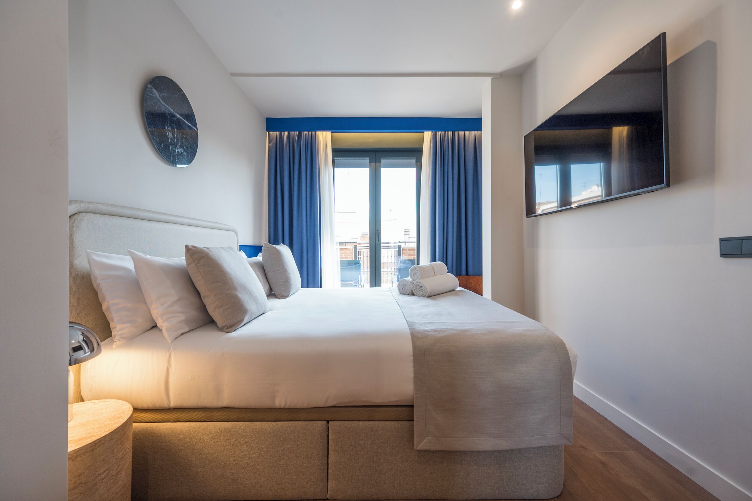 Una habitación de hotel moderna con una cama grande, un televisor de pantalla plana, un reloj de pared redondo y vistas de la ciudad desde la ventana.