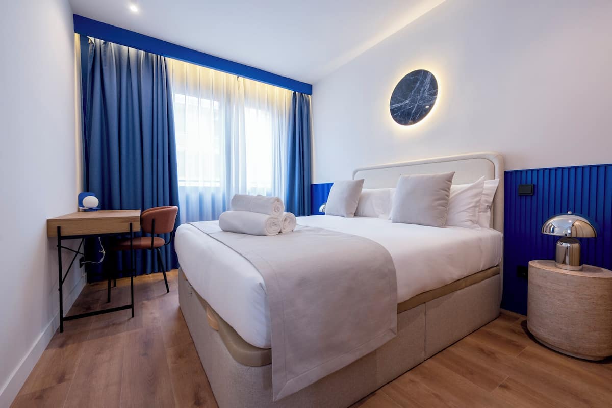 A modern hotel room with a neatly made bed, a desk with a chair, and a round wall clock.