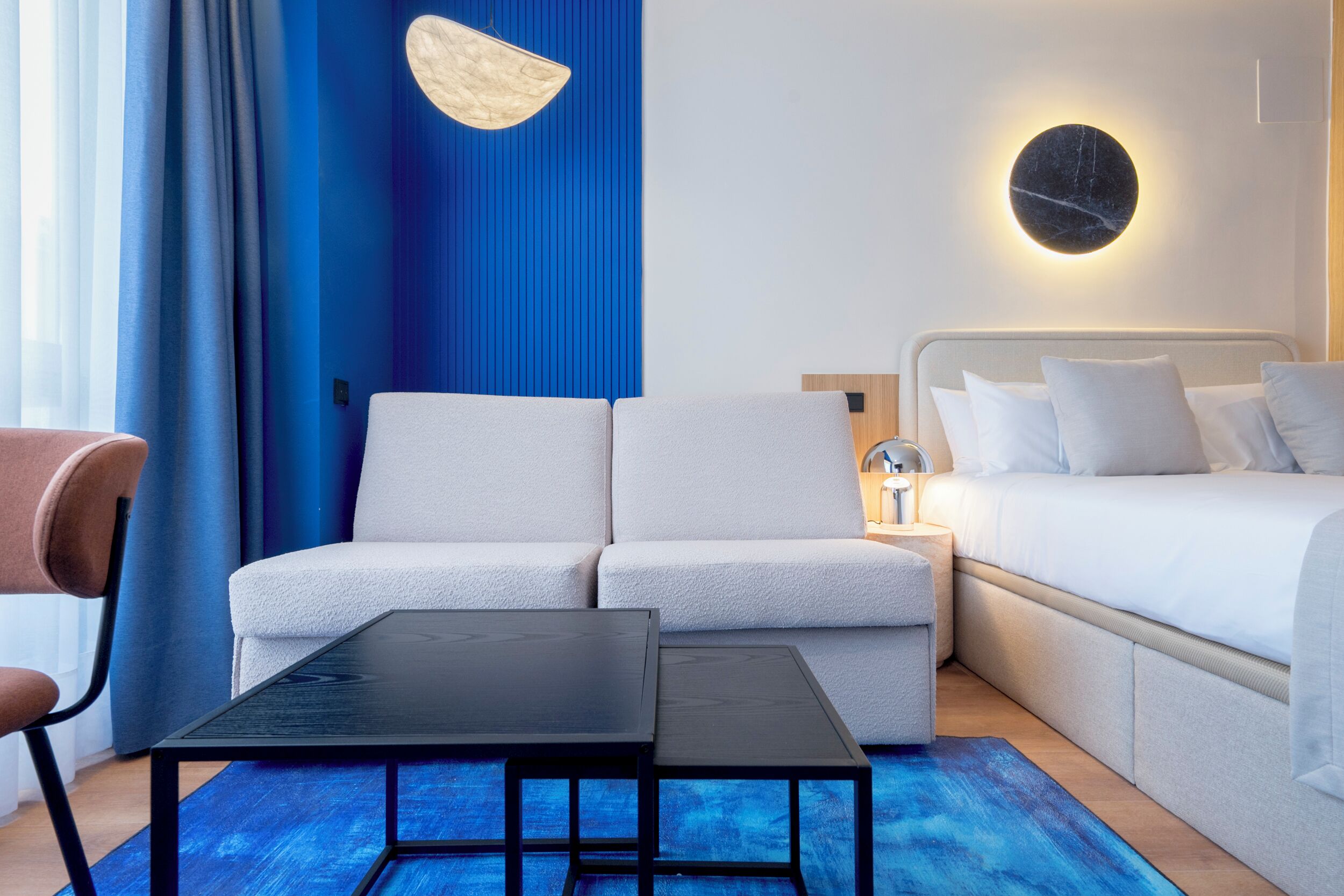 A modern hotel room with a sofa, a bed, a small table, and a wall-mounted light.