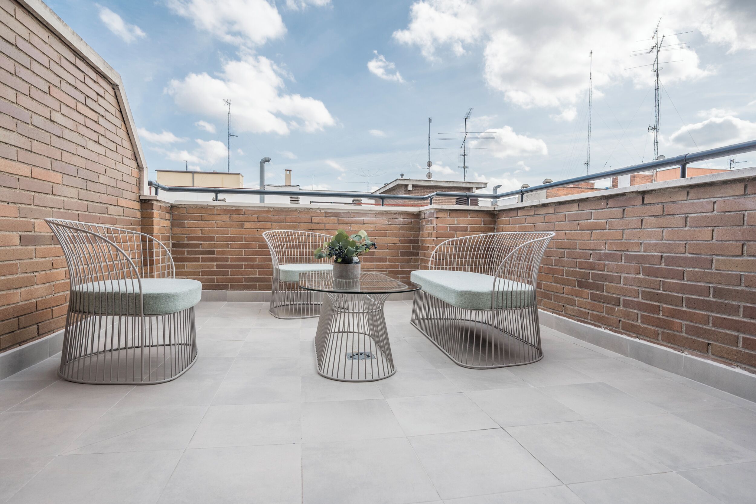 Um terraço no telhado com paredes de tijolo, cadeiras de metal, uma mesa redonda e um vaso de planta.