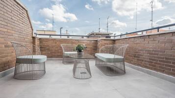 Um terraço no telhado com paredes de tijolo, cadeiras de metal, uma mesa redonda e um vaso de planta.