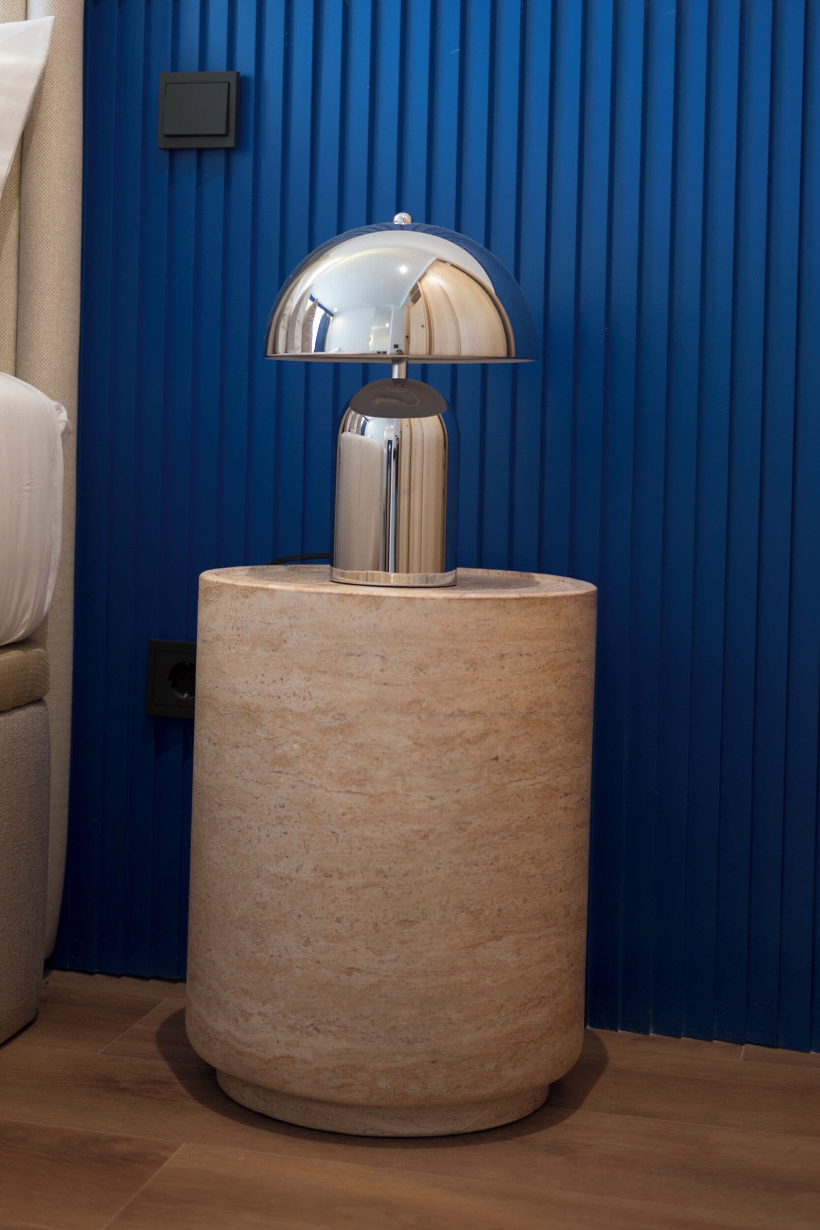 A round, beige side table with a reflective, dome-shaped lamp on top, positioned against a blue vertical panel wall.