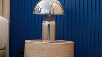A round, beige side table with a reflective, dome-shaped lamp on top, positioned against a blue vertical panel wall.