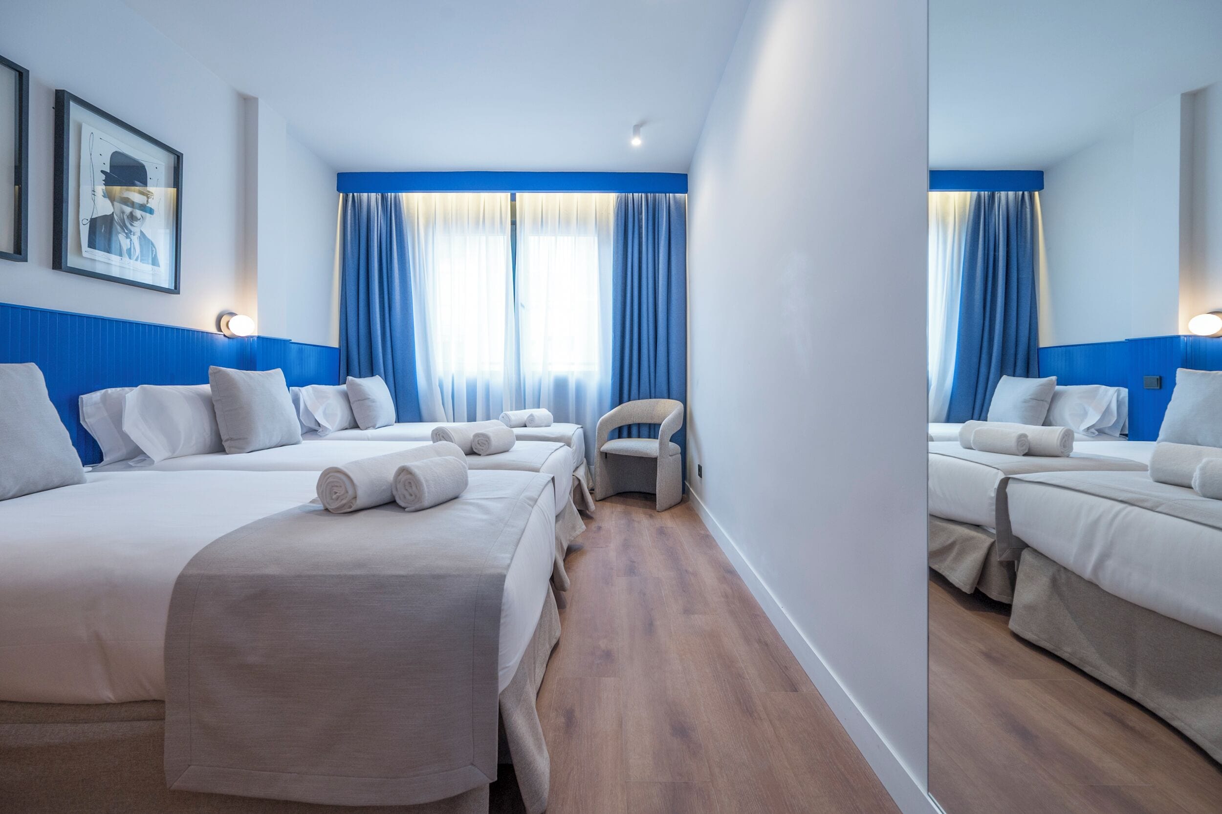A hotel room with two beds, a chair, and a large window with blue curtains.