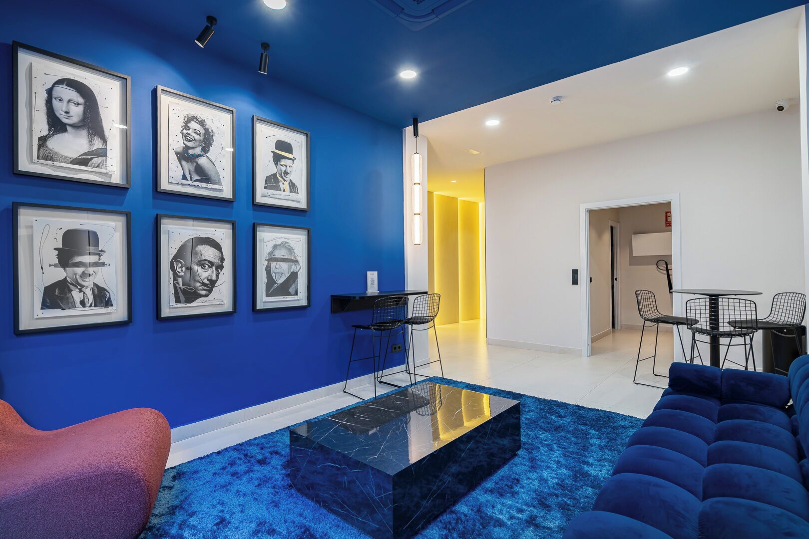 A modern living room with a blue accent wall, a black coffee table, a blue sofa, and a dining area with a small table and chairs.
