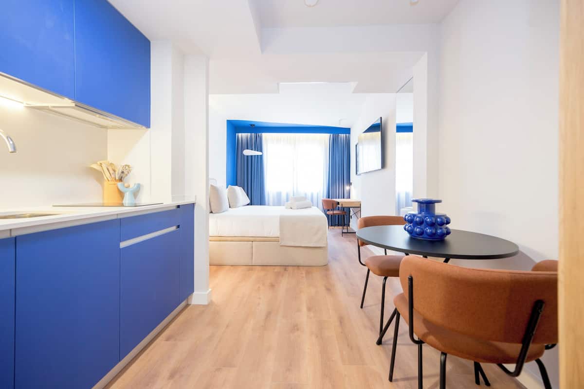 A modern hotel room with a kitchenette, a dining area with a round table and chairs, a bed with a white headboard, and a view of the city through large windows.