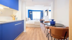 Quarto de hotel moderno com cozinha compacta, área de jantar com mesa redonda e cadeiras, cama com cabeceira branca e vista da cidade através de amplas janelas.