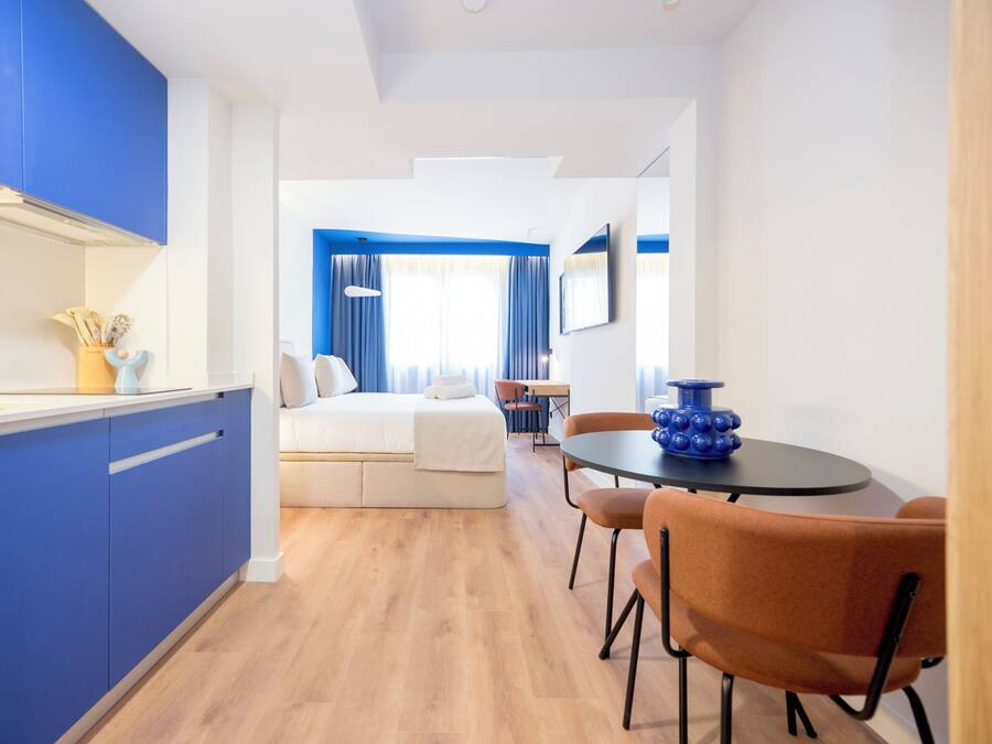 A modern hotel room with a kitchenette, a dining area with a round table and chairs, a bed with a white headboard, and a view of the city through large windows.