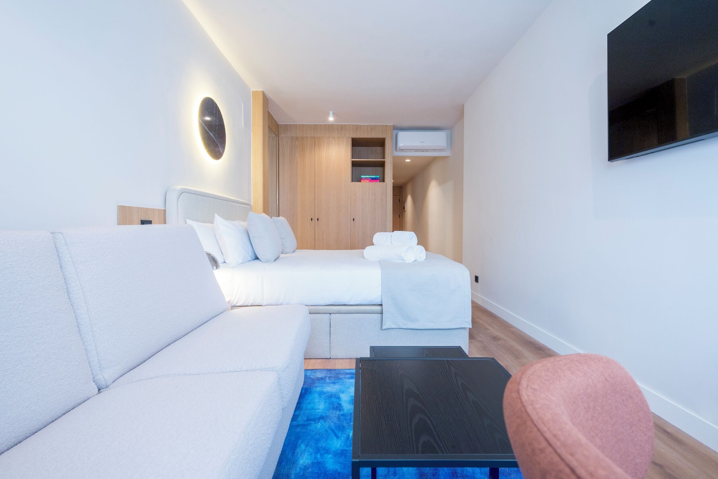 Quarto de hotel moderno com um sofá branco, uma cama com roupa de cama branca, uma mesa pequena preta e uma TV fixada na parede.