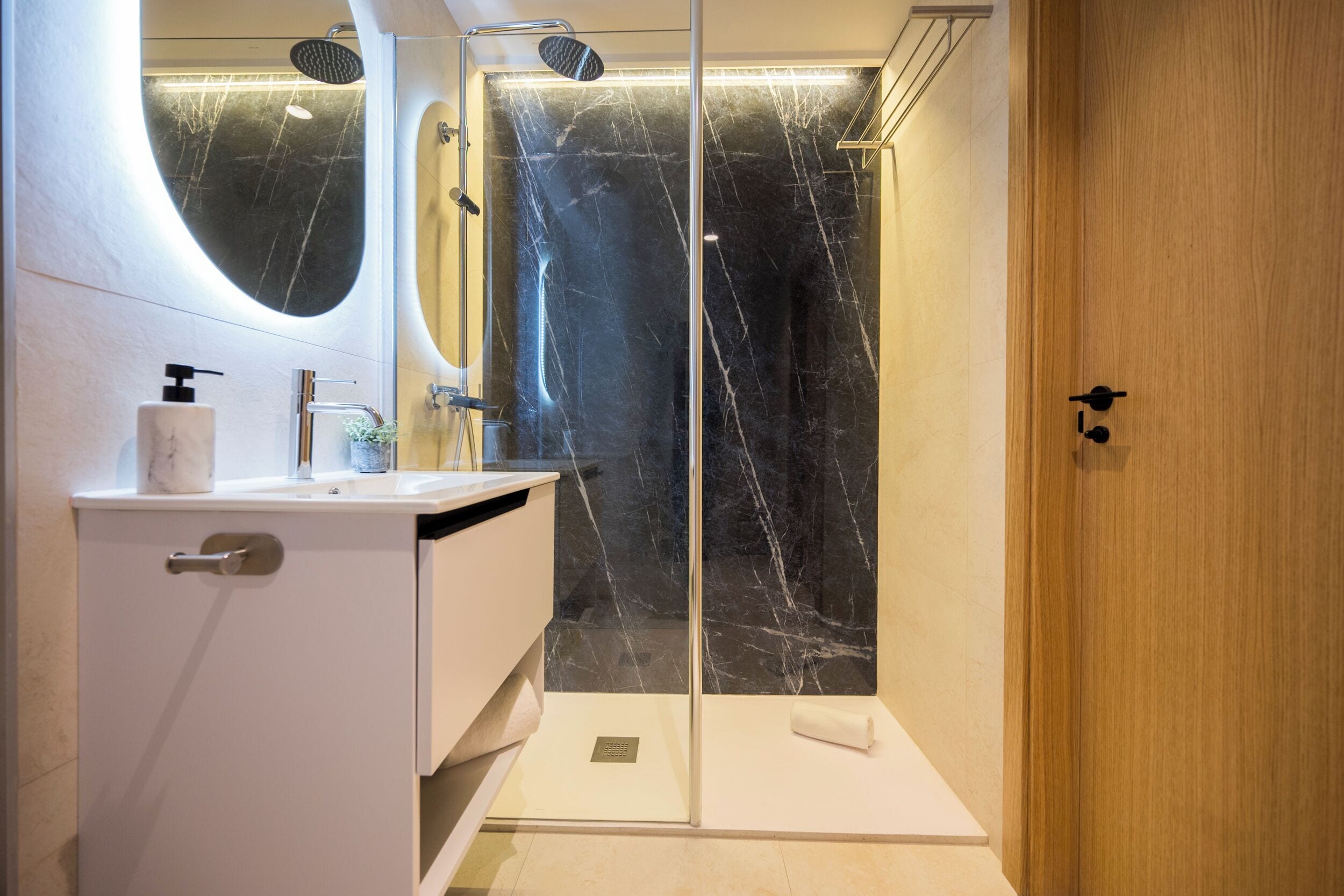A modern bathroom with a marble shower enclosure, a white vanity with a large mirror, and a wooden door.