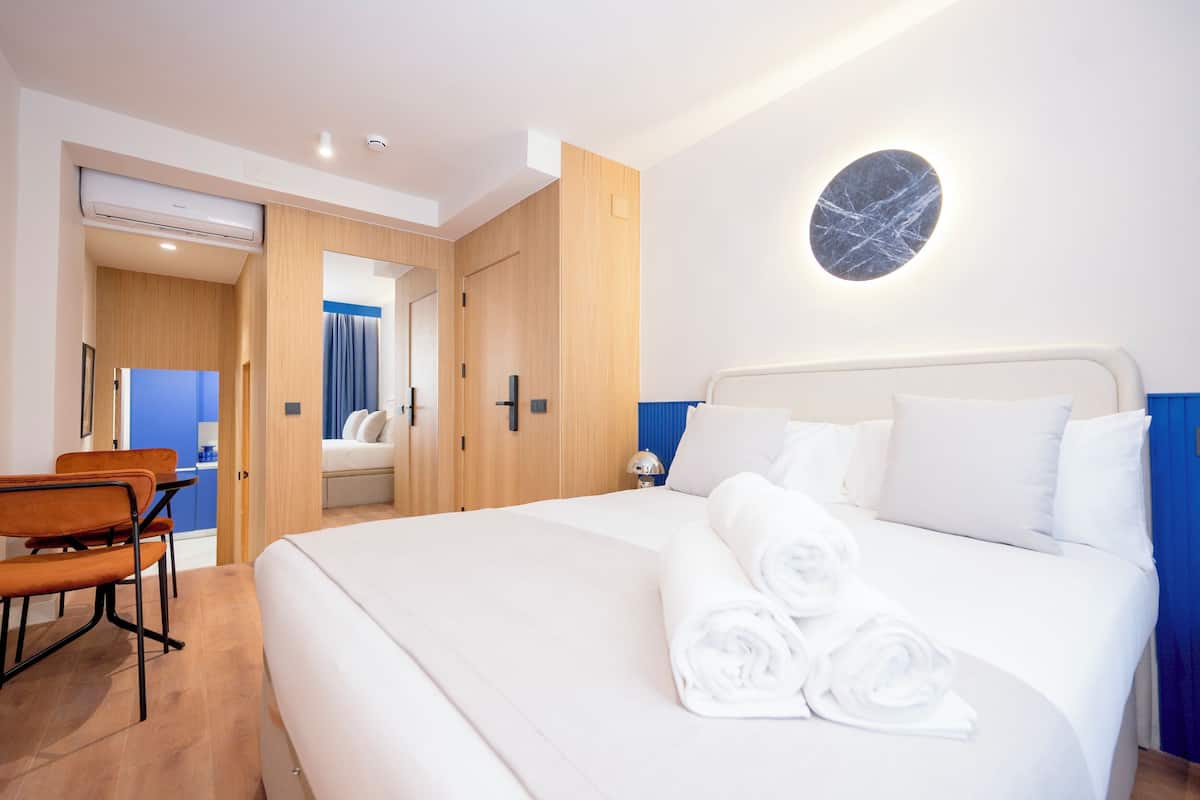 A hotel room with a large bed, a desk, and a chair. There is a wall-mounted light fixture above the bed.