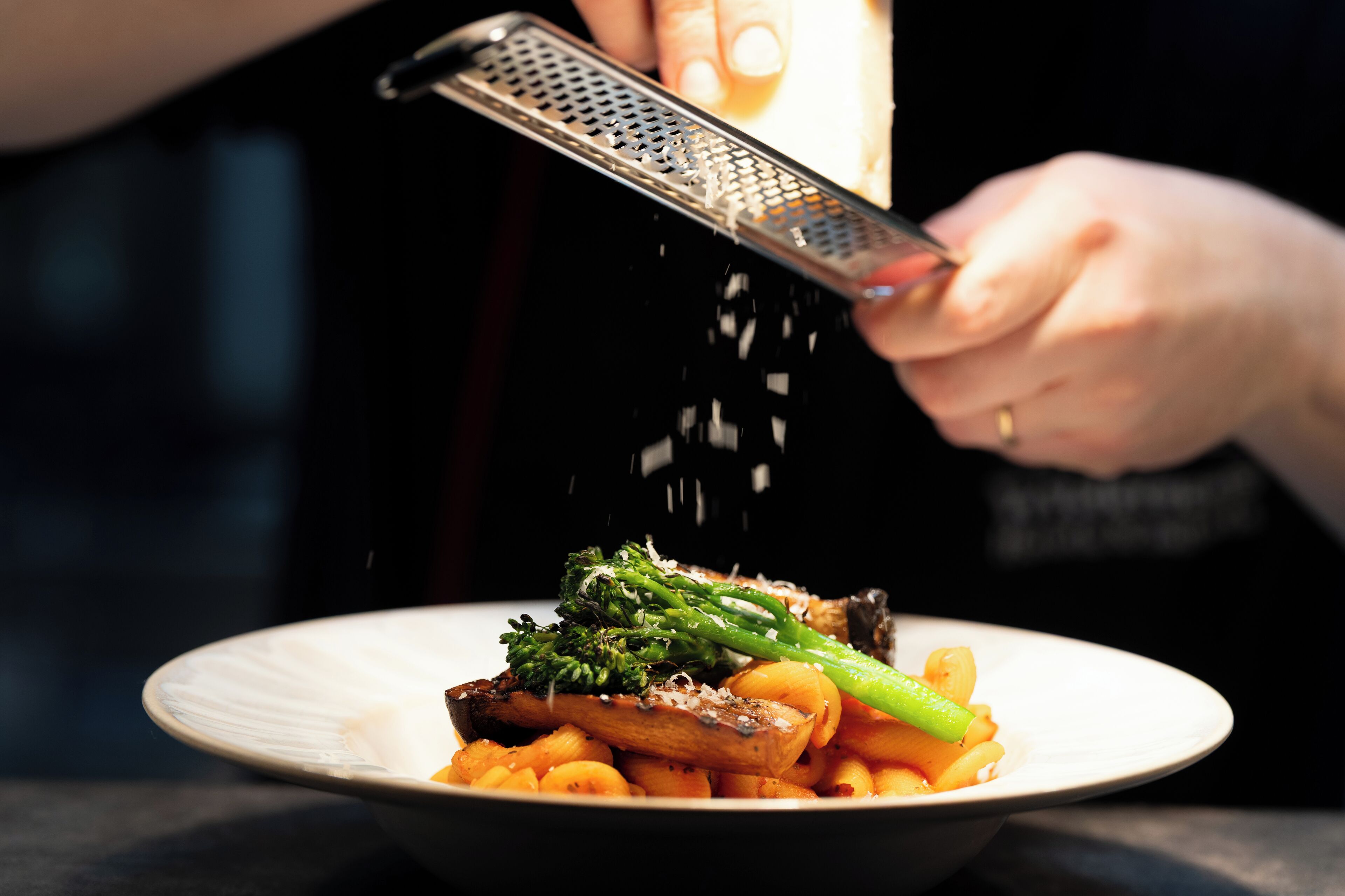 A person grating cheese over a dish of pasta and vegetables.