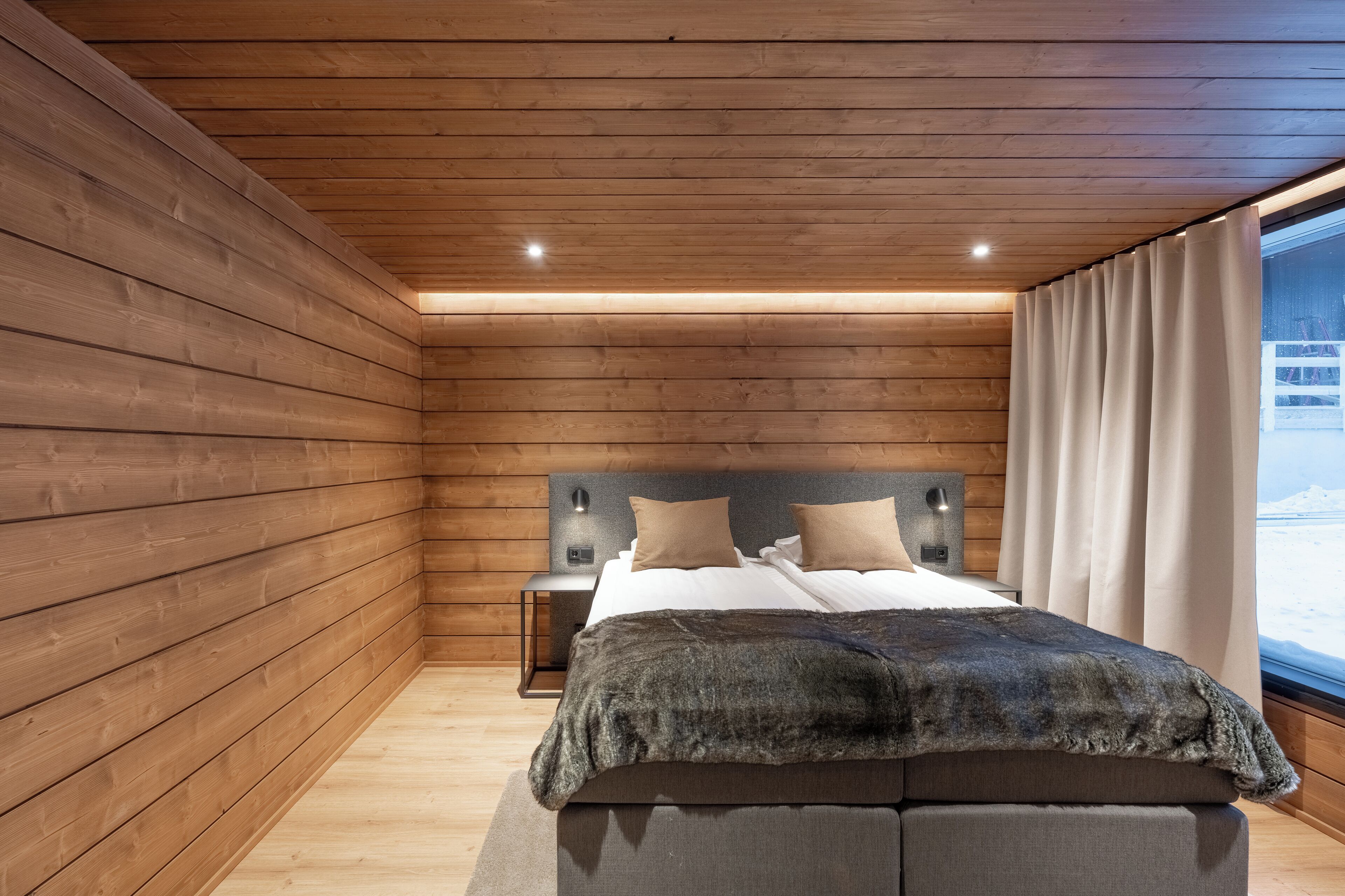 A cozy bedroom with wooden walls, a bed with a dark blanket, two pillows, and a nightstand with a lamp.