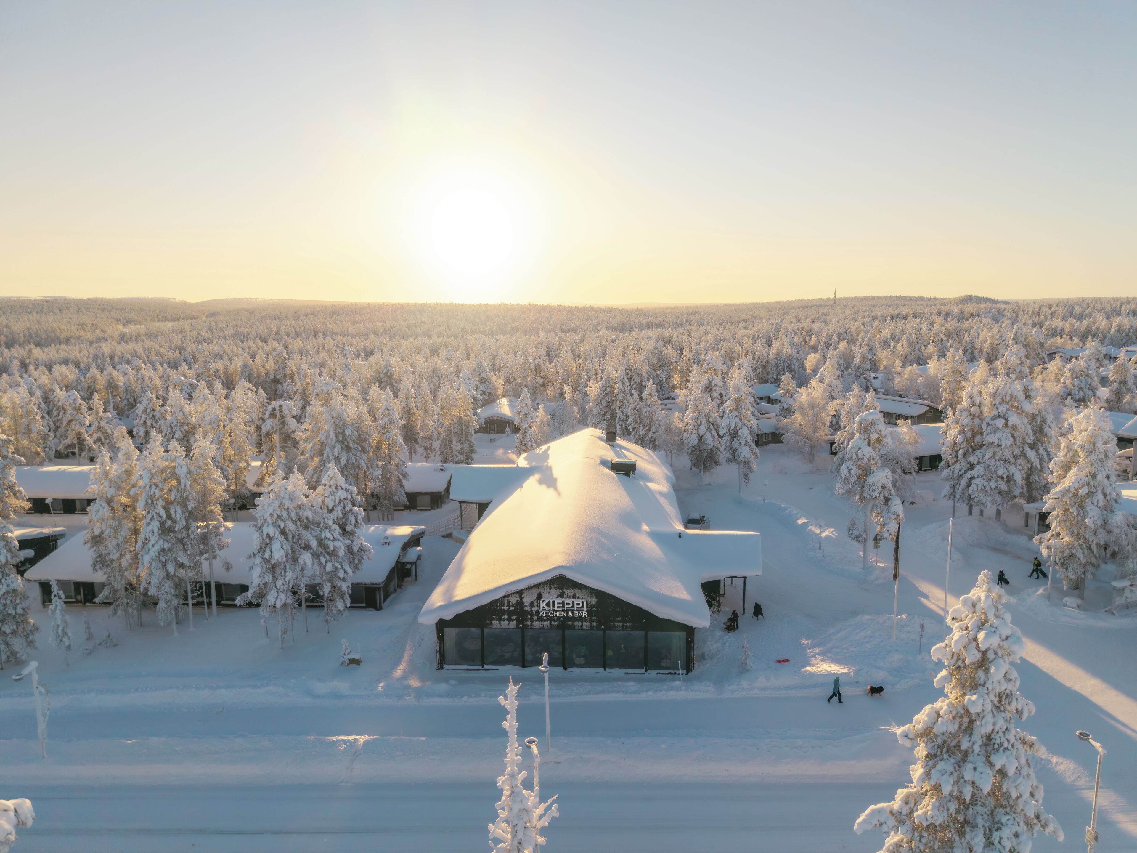 A snow-covered landscape with buildings and trees, including a prominent building with a sign that reads "Kieppi Kitchen Bar".