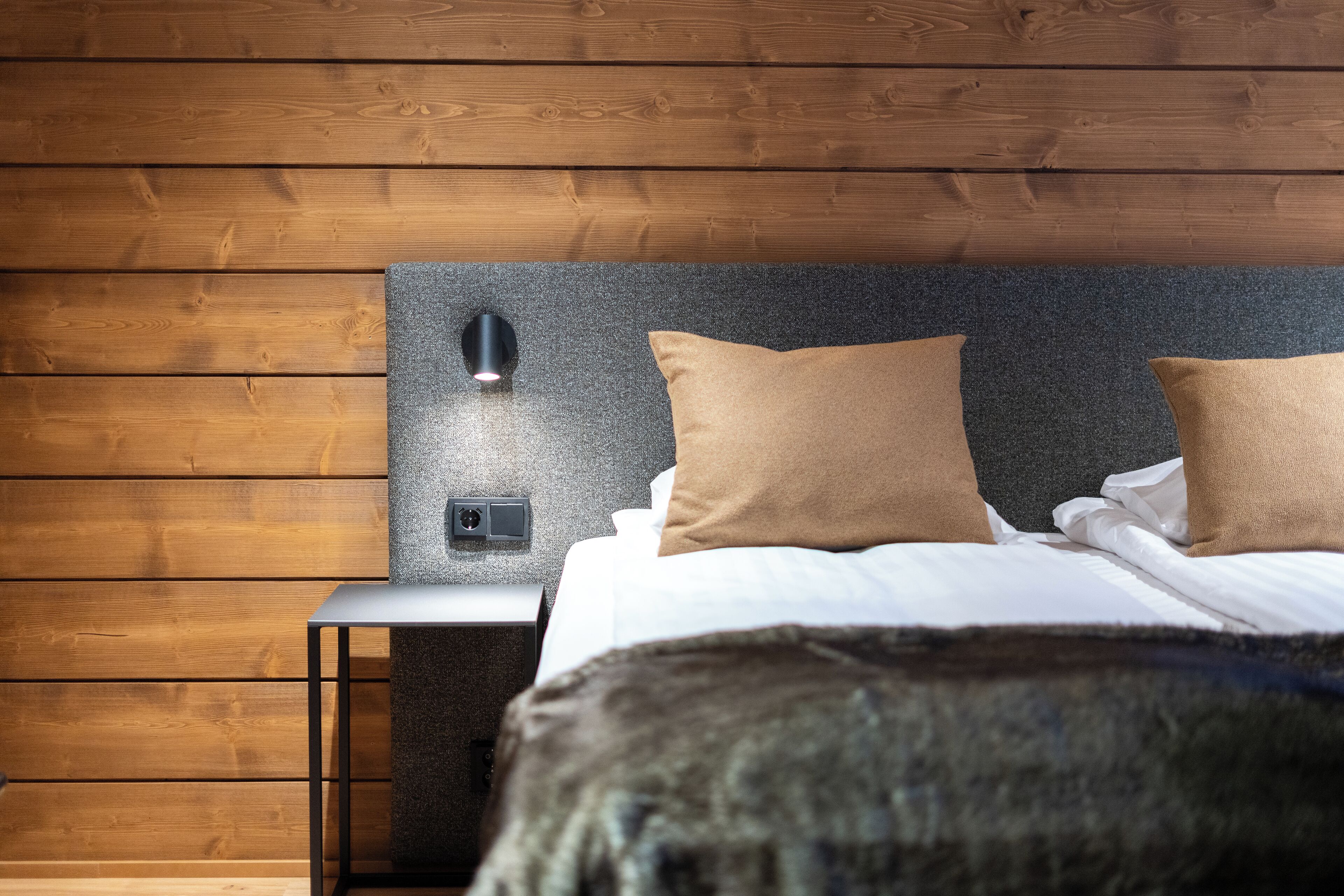 Ein ordentlich bezogenes Bett mit zwei Kissen, ein Nachttisch mit Lampe sowie eine Wand mit Holzvertäfelung.