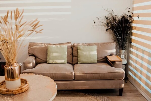 A beige leather sofa with two cushions, a woven side table, a vase with dried plants, and a striped wall in the background.