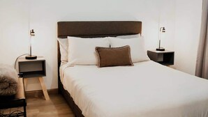 Una cama bien hecha con un cabecero marrón, dos mesitas de noche con lámparas y un pequeño taburete de madera.