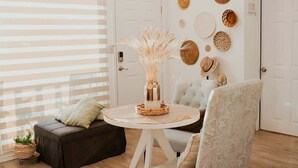 Una sala de estar acogedora con una mesa redonda blanca, un sofá marrón oscuro y un sillón estampado. Un jarrón decorativo con plantas secas está sobre la mesa.