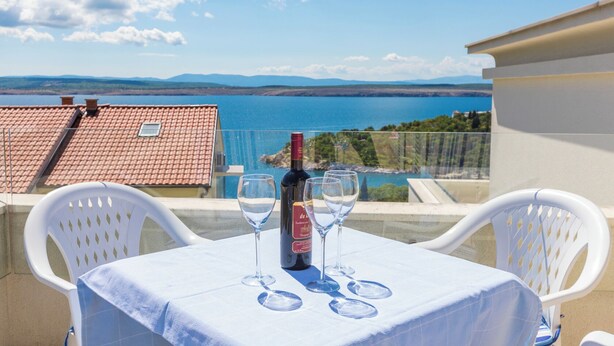 A table with a bottle of wine and glasses on a balcony overlooking a scenic view of a body of water and distant mountains. - Apartment Mirta 1 (Dramalj)