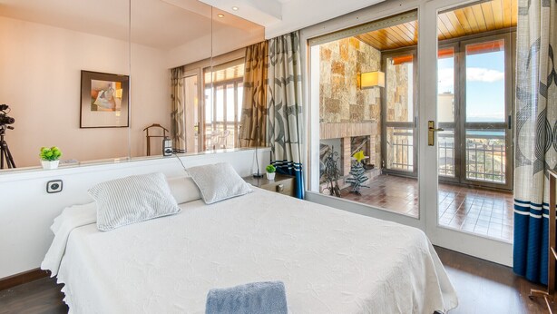 A bedroom with a bed, a desk, a chair, a potted plant, and a view of the outside.