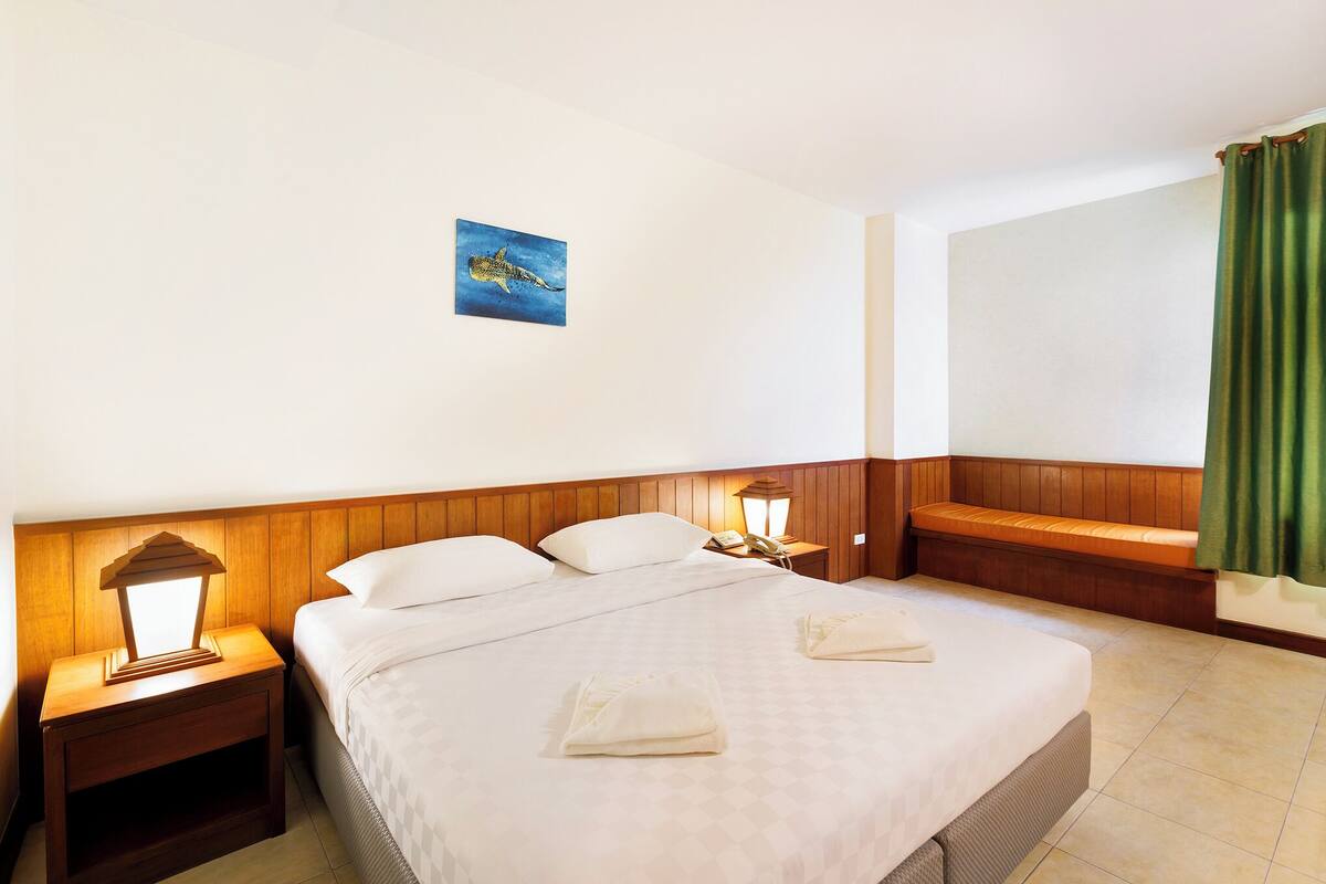 Ein Hotelzimmer mit einem großen Bett, einem hölzernen Kopfteil, zwei Nachttischen mit Lampen, einem kleinen Gemälde an der Wand und einem grünen Vorhang.