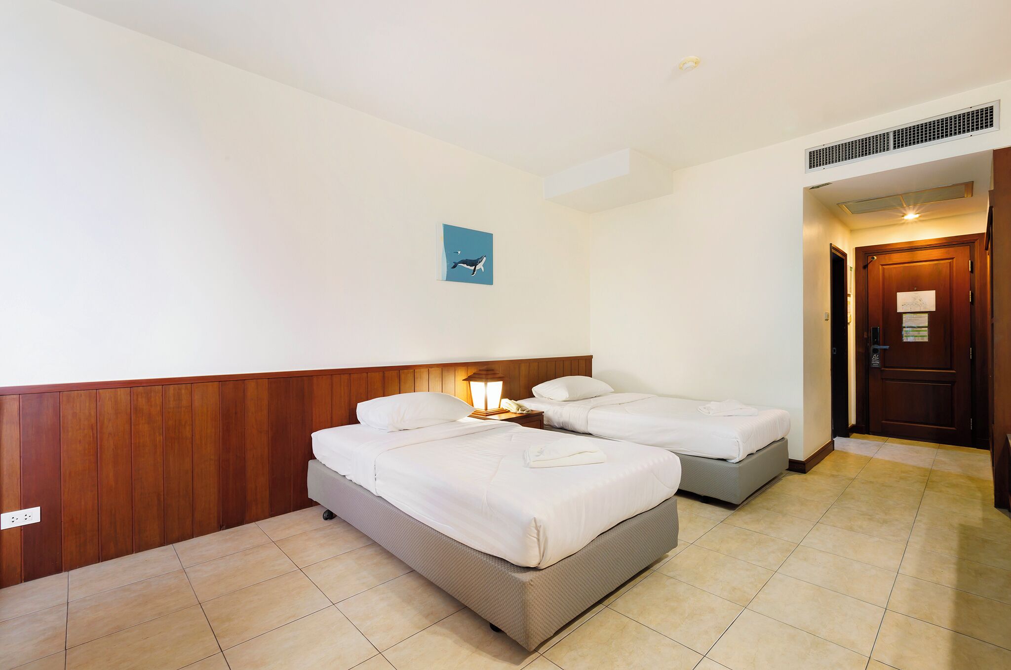 A hotel room with two single beds, a wooden headboard, a nightstand with a lamp, and a small wall art.