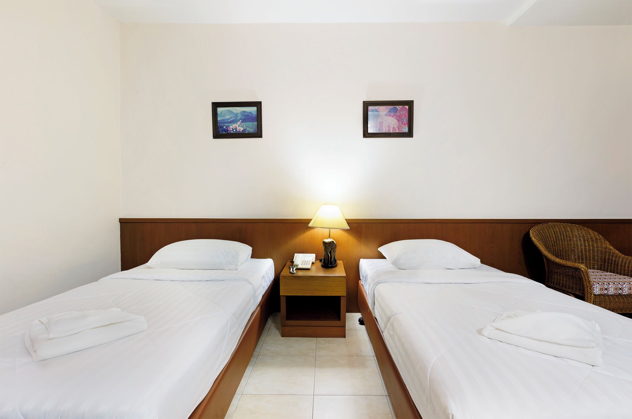 A hotel room with two single beds, a nightstand with a lamp, and two framed pictures on the wall.