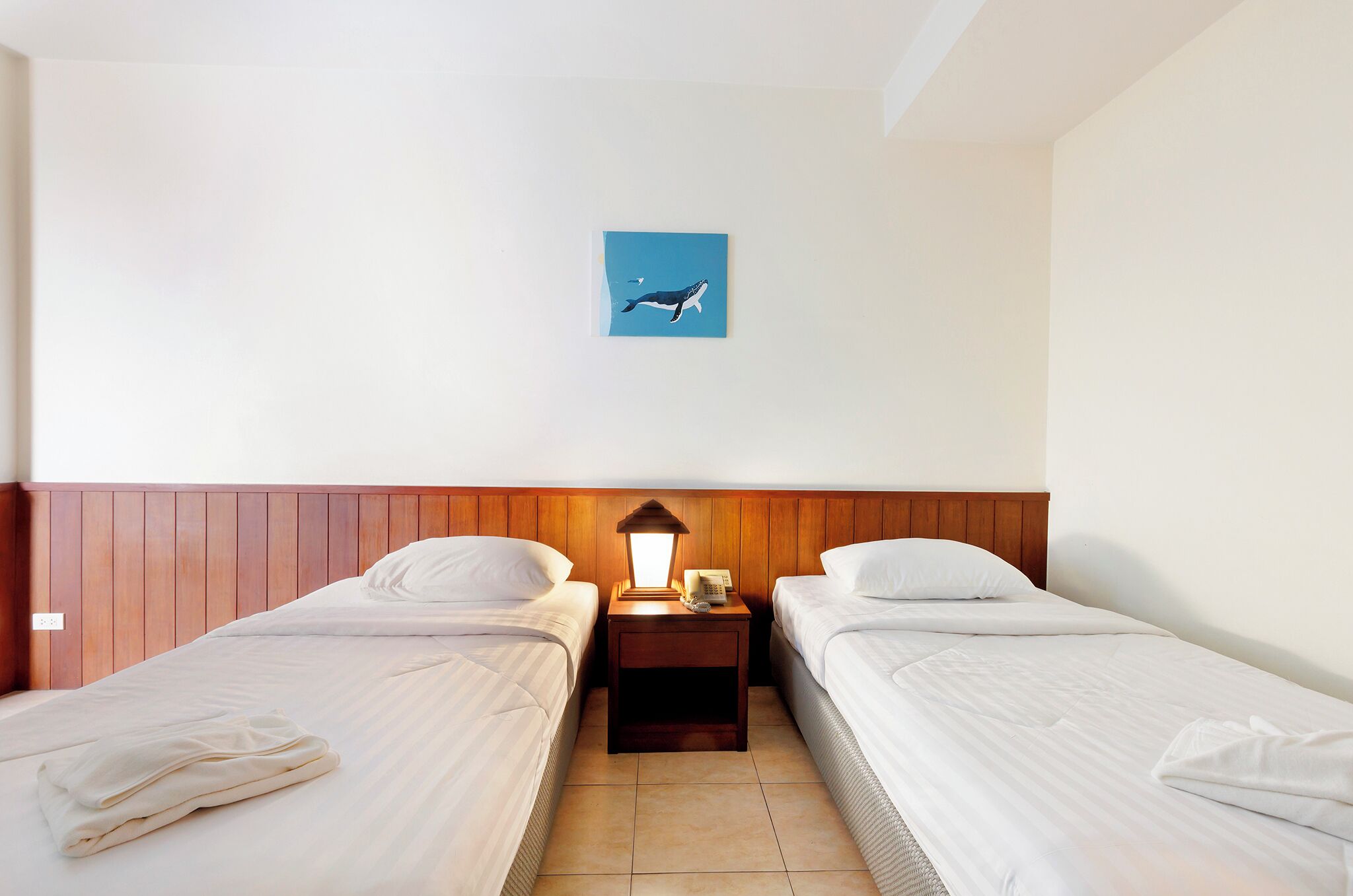 Ein Hotelzimmer mit zwei Einzelbetten, einem hölzernen Kopfteil, einem Nachttisch mit Lampe und einem an der Wand befestigten Bild von einem Flugzeug.