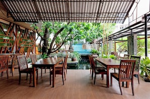 A covered outdoor dining area with wooden tables and chairs, surrounded by greenery and a view of a building in the background.