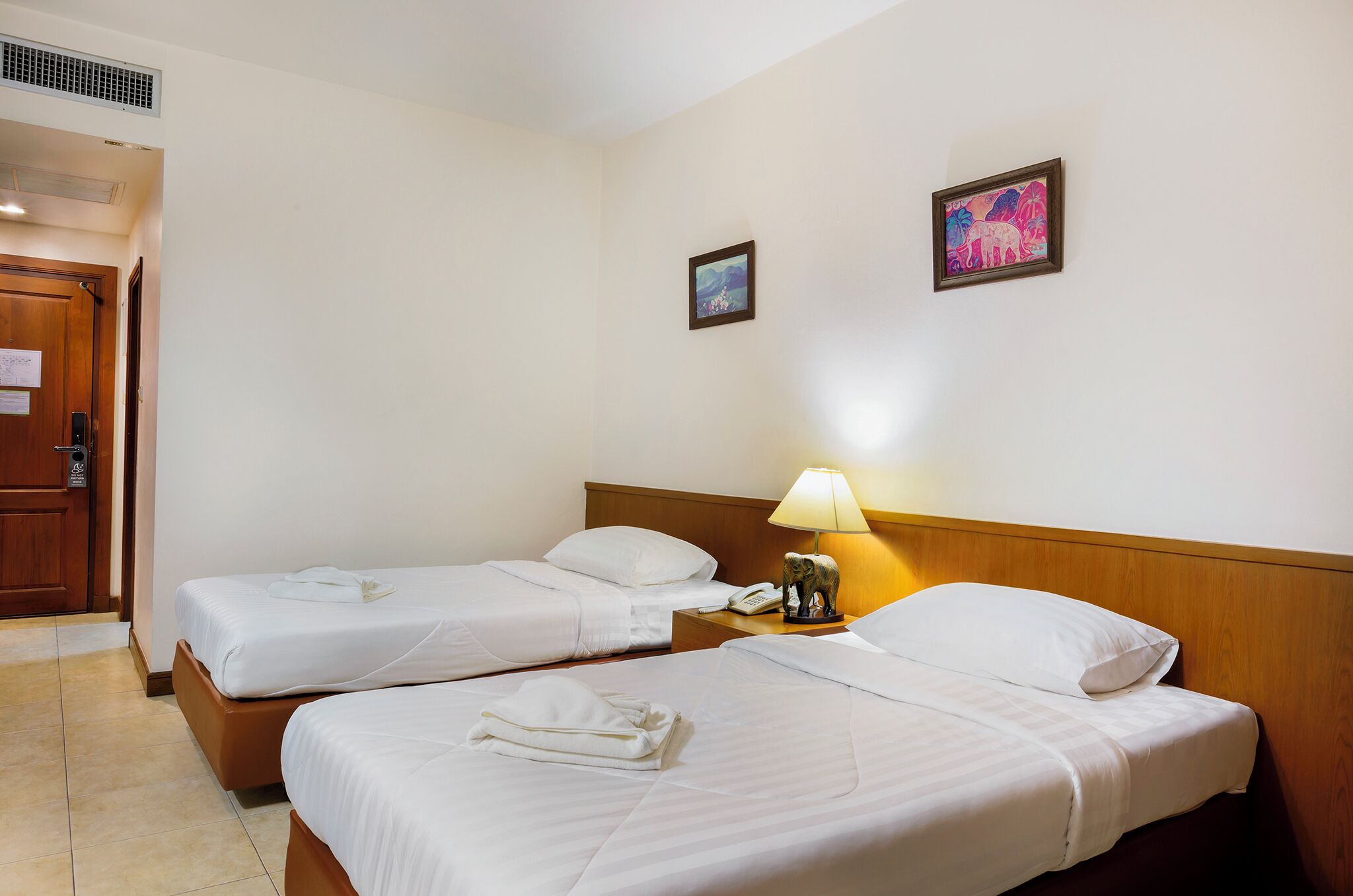 Una habitación de hotel con dos camas individuales, un cabecero de madera, una mesita de noche con una lámpara y un pequeño cuadro en la pared.