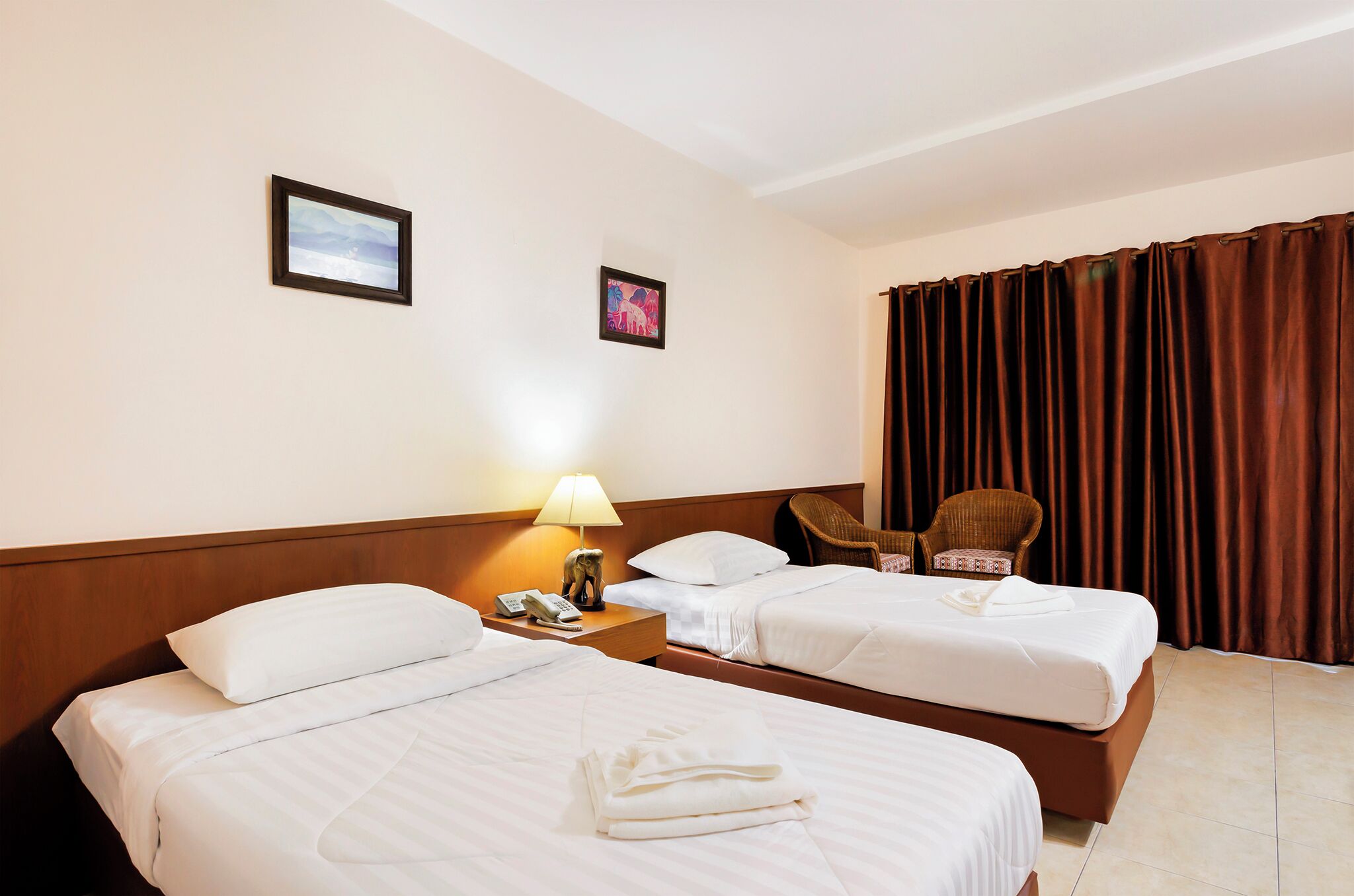 Une chambre d’hôtel avec deux lits, une table de chevet avec une lampe, une chaise et deux tableaux encadrés au mur.