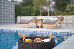 Un petit-déjeuner servi au bord de la piscine, avec un panier en osier contenant un verre de jus d’orange, un verre d’eau, des raisins, un muffin et un bol de céréales.