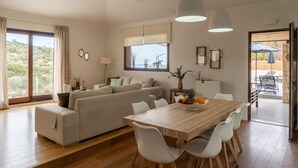A modern living room with a sofa, dining table, and chairs. There is a window with curtains, a lamp, and a view of the outdoors. - Villa in Kastellákia with Private Pool & BBQ (Rethymno)