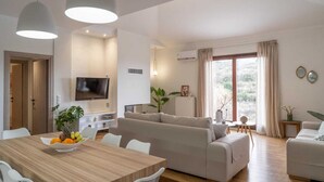 A modern living room with a sofa, a dining table with fruit, and a TV. - Villa in Kastellákia with Private Pool & BBQ (Rethymno)