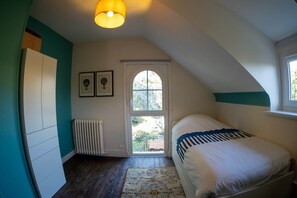 Een slaapkamer met een schuine plafond, een radiator, een bed met een gestreept dekbed, een raam met uitzicht op groen, en een deur die naar buiten leidt.