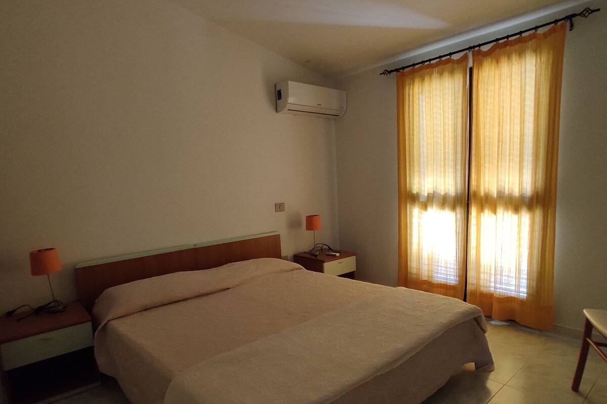 A hotel room with a large bed, two bedside tables, an air conditioning unit, and a window with curtains.