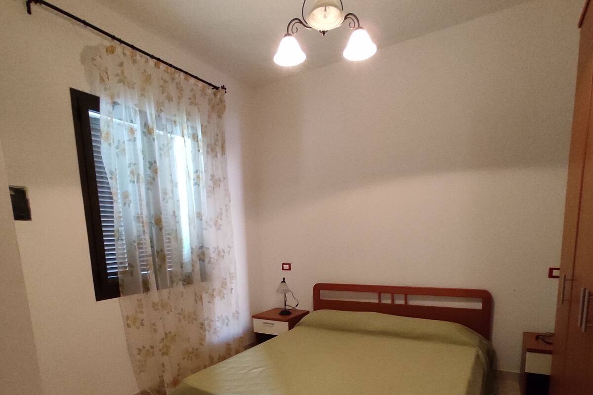 A bedroom with a bed, a nightstand, a lamp, a window with curtains, and a wooden wardrobe.