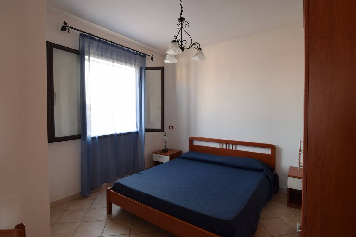 A bedroom with a bed, a nightstand, a lamp, a window with curtains, and a ceiling light fixture.