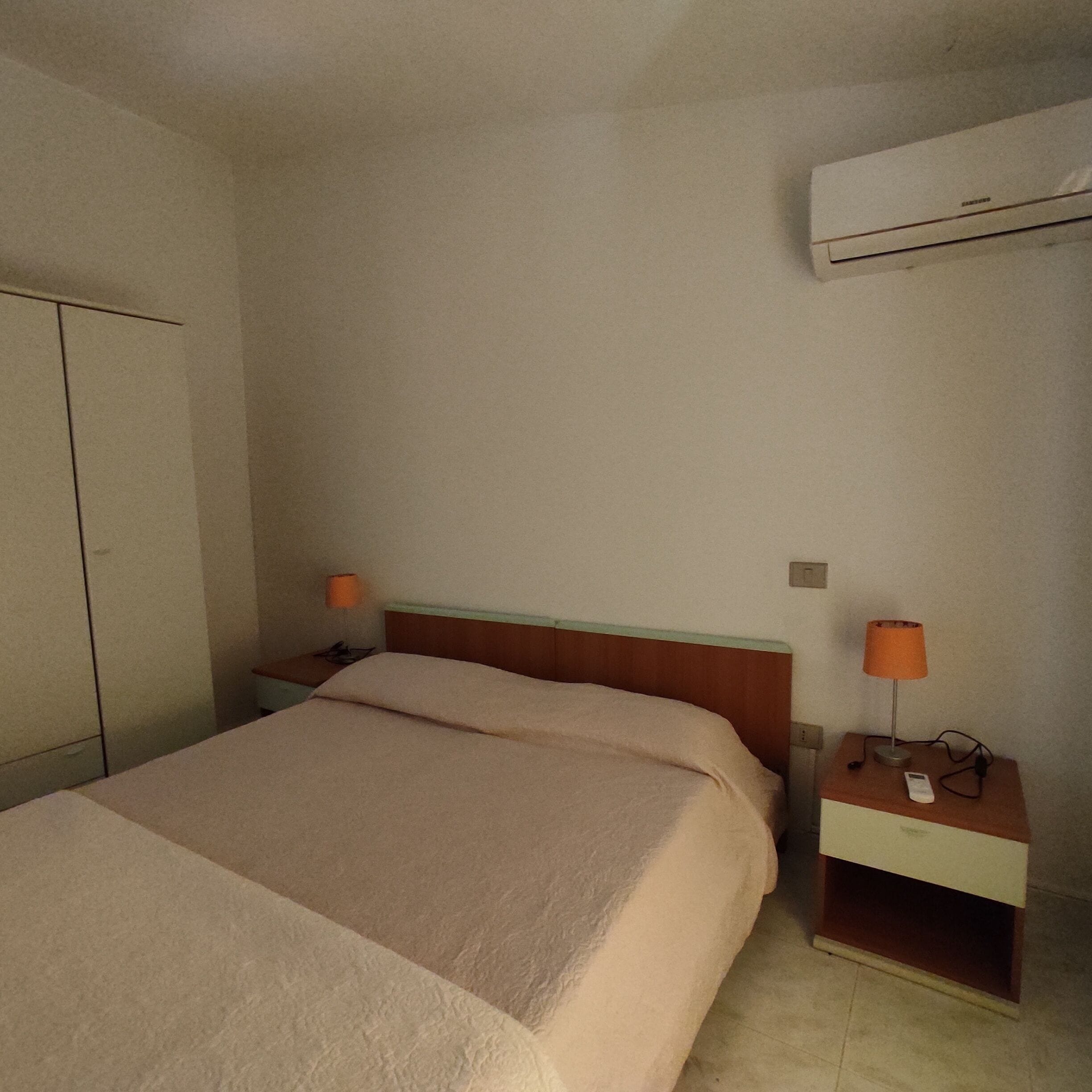 A simple, clean hotel room with a bed, a nightstand with a lamp, a wardrobe, and an air conditioning unit on the wall.